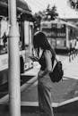 Black and white photo of a woman at a city bus stop, using her phone and wearing casual attire.